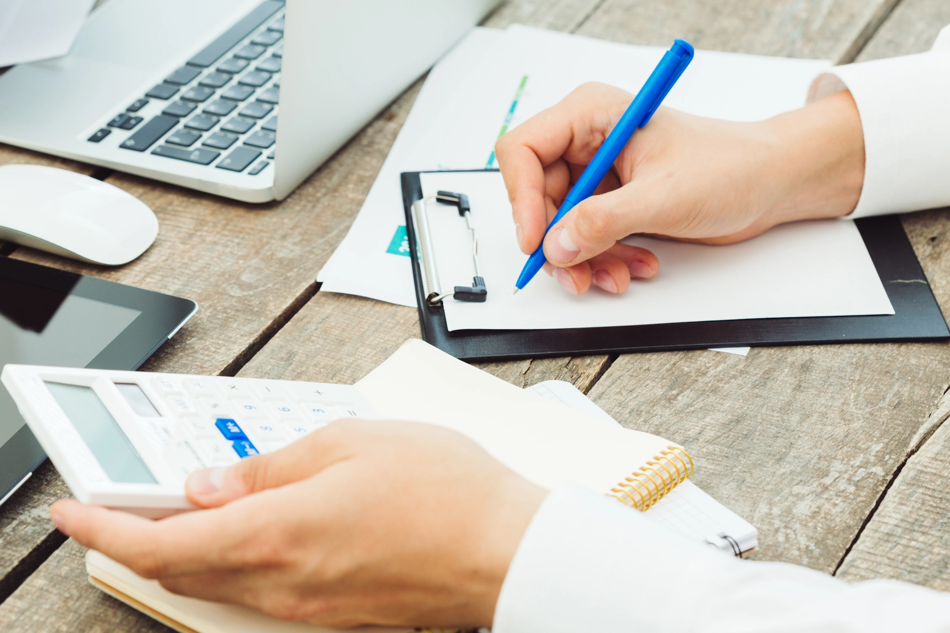 A hand writing on a clipboard with a pen, alongside a laptop, calculator and notebook,