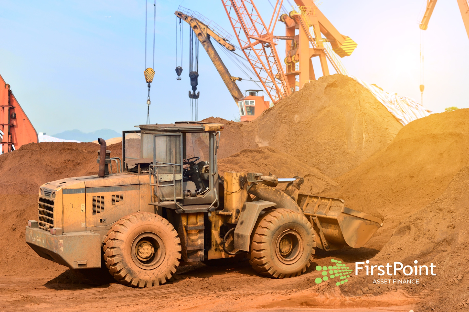 Loader machinery operating on a construction site with piles of sand and cranes in the